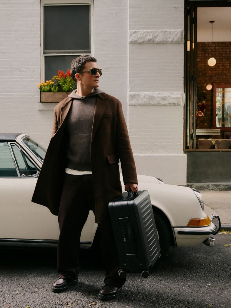 Man in a brown coat wearing sunglasses holds a black hard-shell suitcase next to a white classic car in the city.