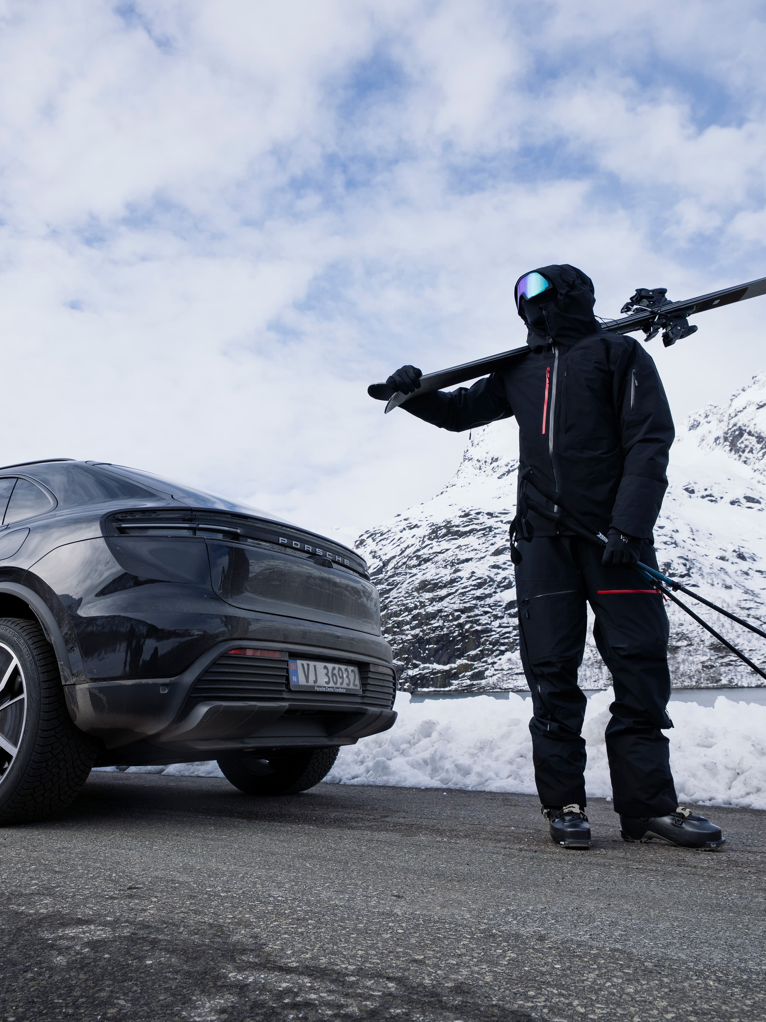 Person in schwarzer Skibekleidung mit Skiern auf der Schulter steht neben einem Porsche in verschneiter Berglandschaft.