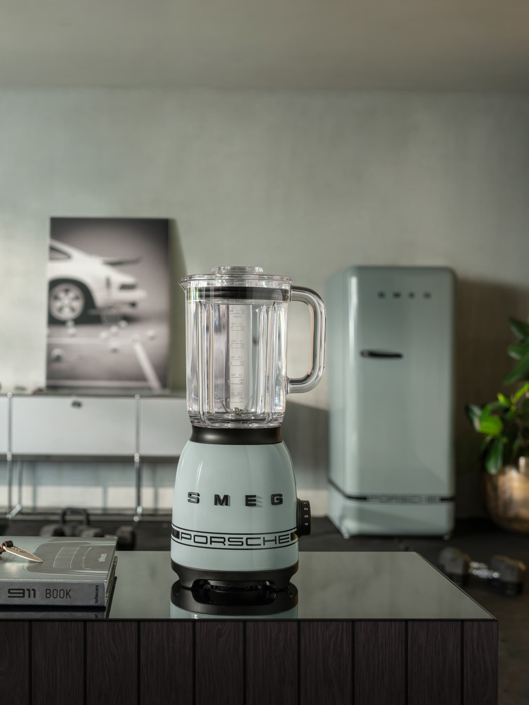 Pale blue Smeg Porsche blender on a dark counter, with a vintage fridge and car poster in the background.