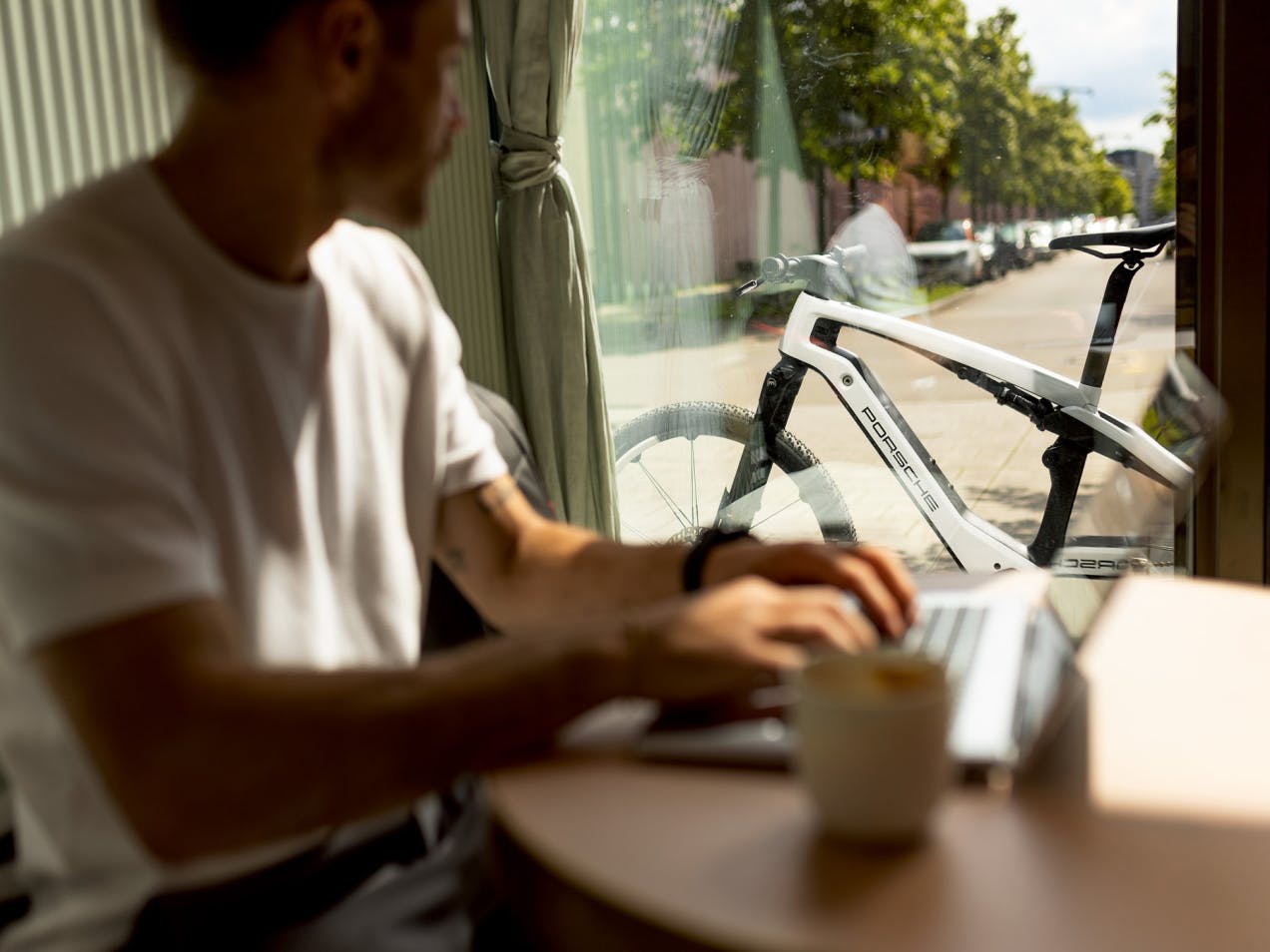 A man sits at his laptop in a cafe and in the background is the new Porsche Ebike Sport in white.