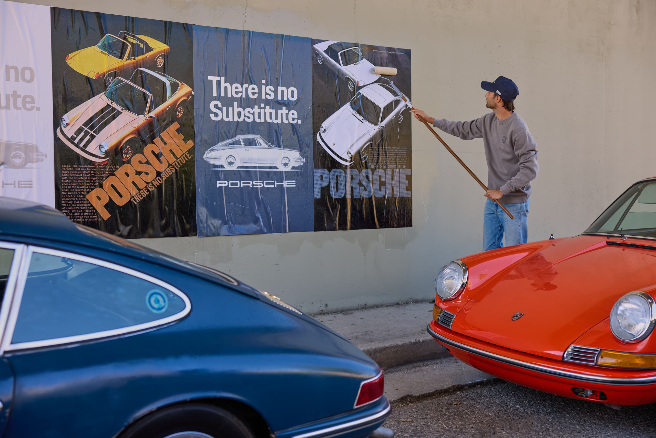 Man applying a Porsche poster to a wall with a paint roller, with blue and red classic Porsche cars parked in the foreground.