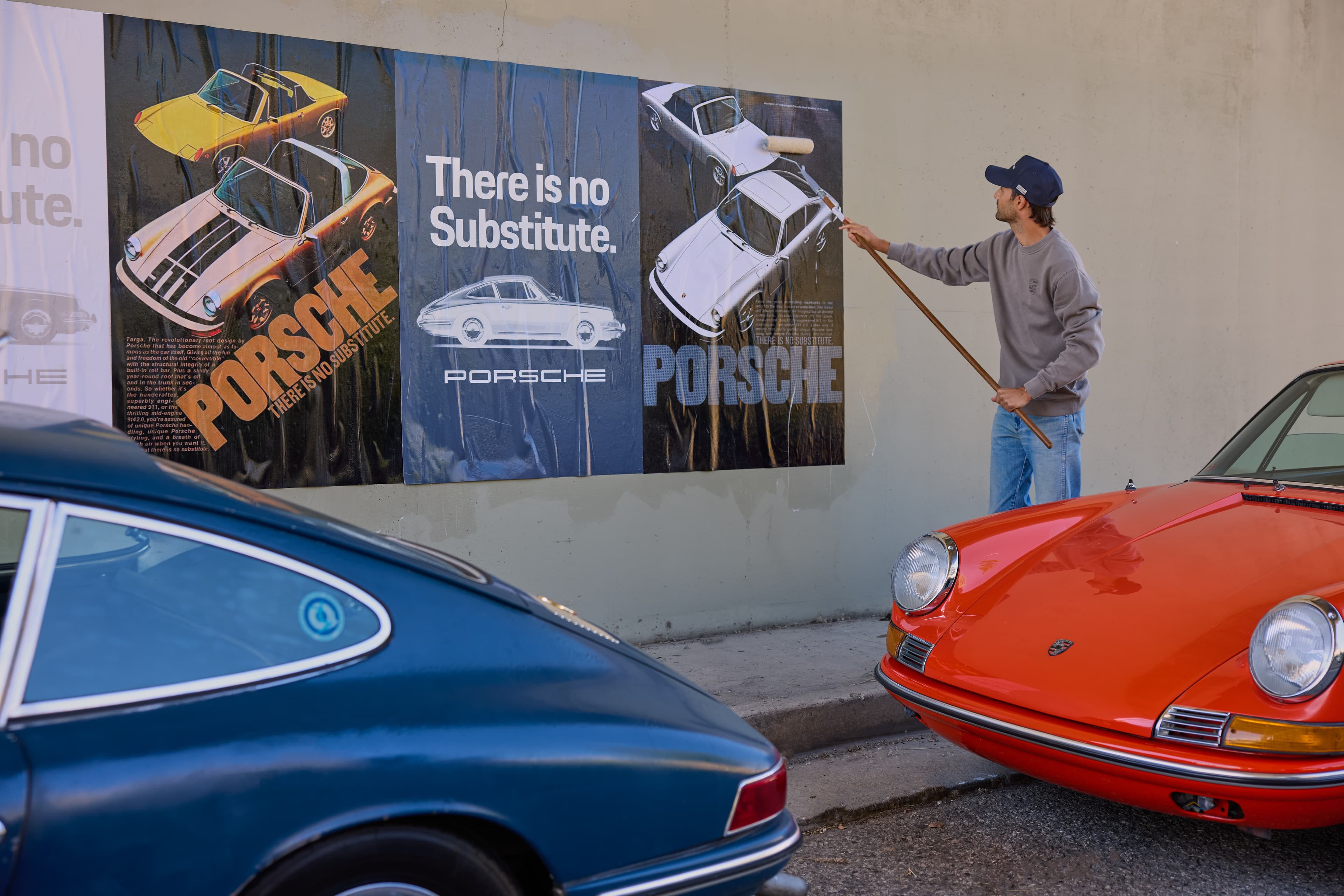 Man applying a Porsche poster to a wall with a paint roller, with blue and red classic Porsche cars parked in the foreground.