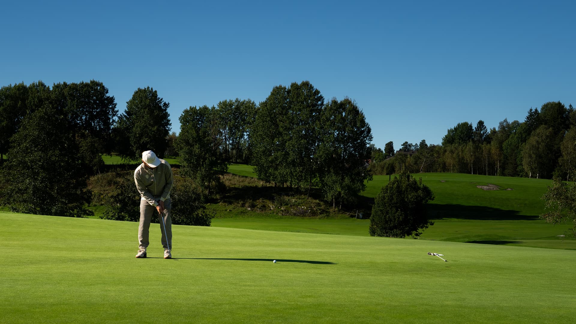 Golfer in checkered outfit putting on a green, with trees and clear blue sky in the background.
