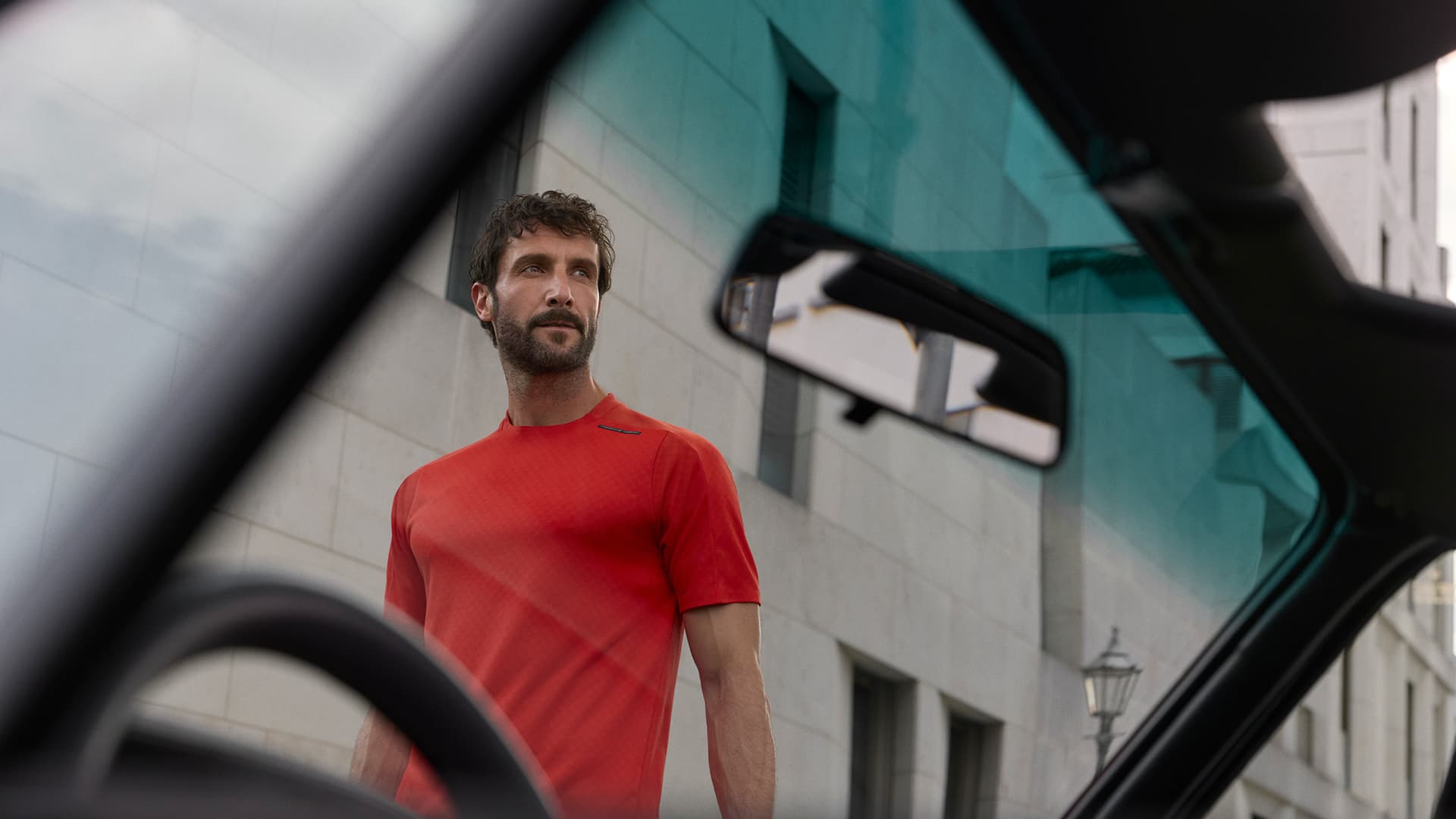 Man in red shirt seen through car window, standing against a modern building with a thoughtful expression.
