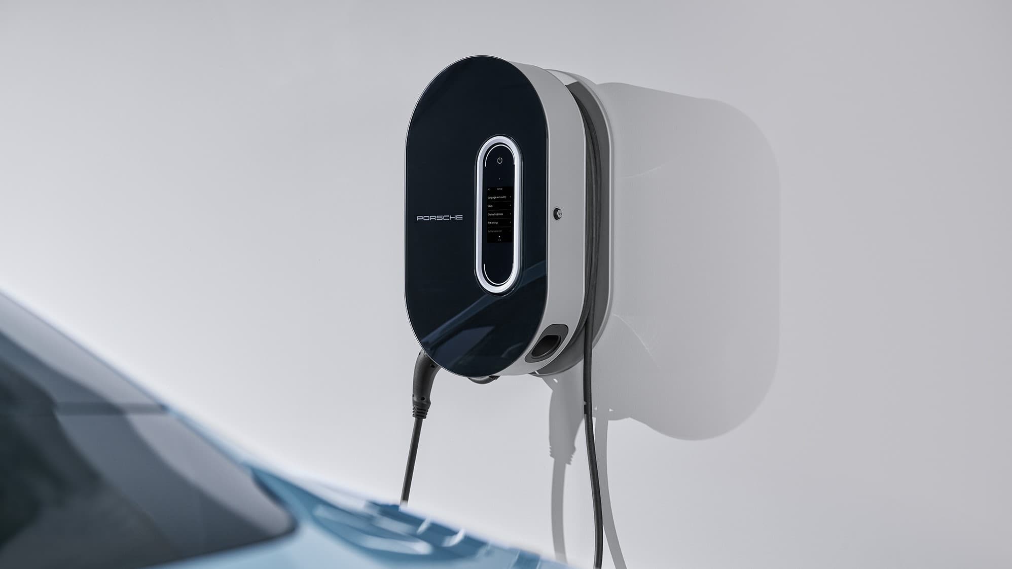Chargeur mural Porsche en noir et blanc, monté sur un mur blanc, avec une voiture bleue partiellement visible au premier plan.