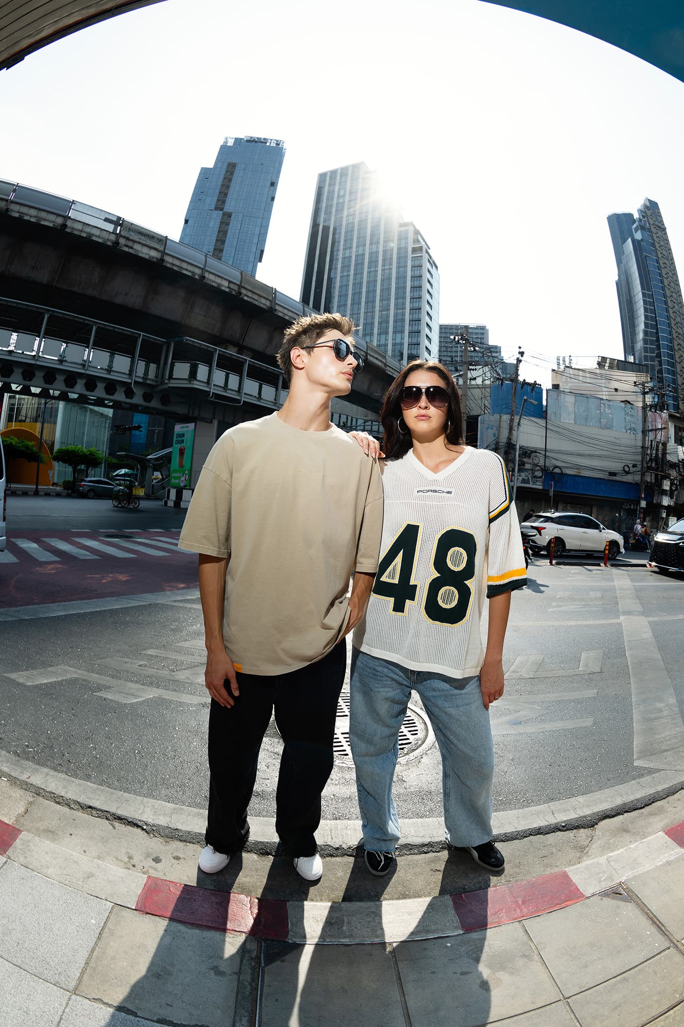 Two models in sunglasses wear the Porsche AHEAD Collection: a beige T-shirt and a white Porsche jersey with number 48, set against tall skyscrapers.