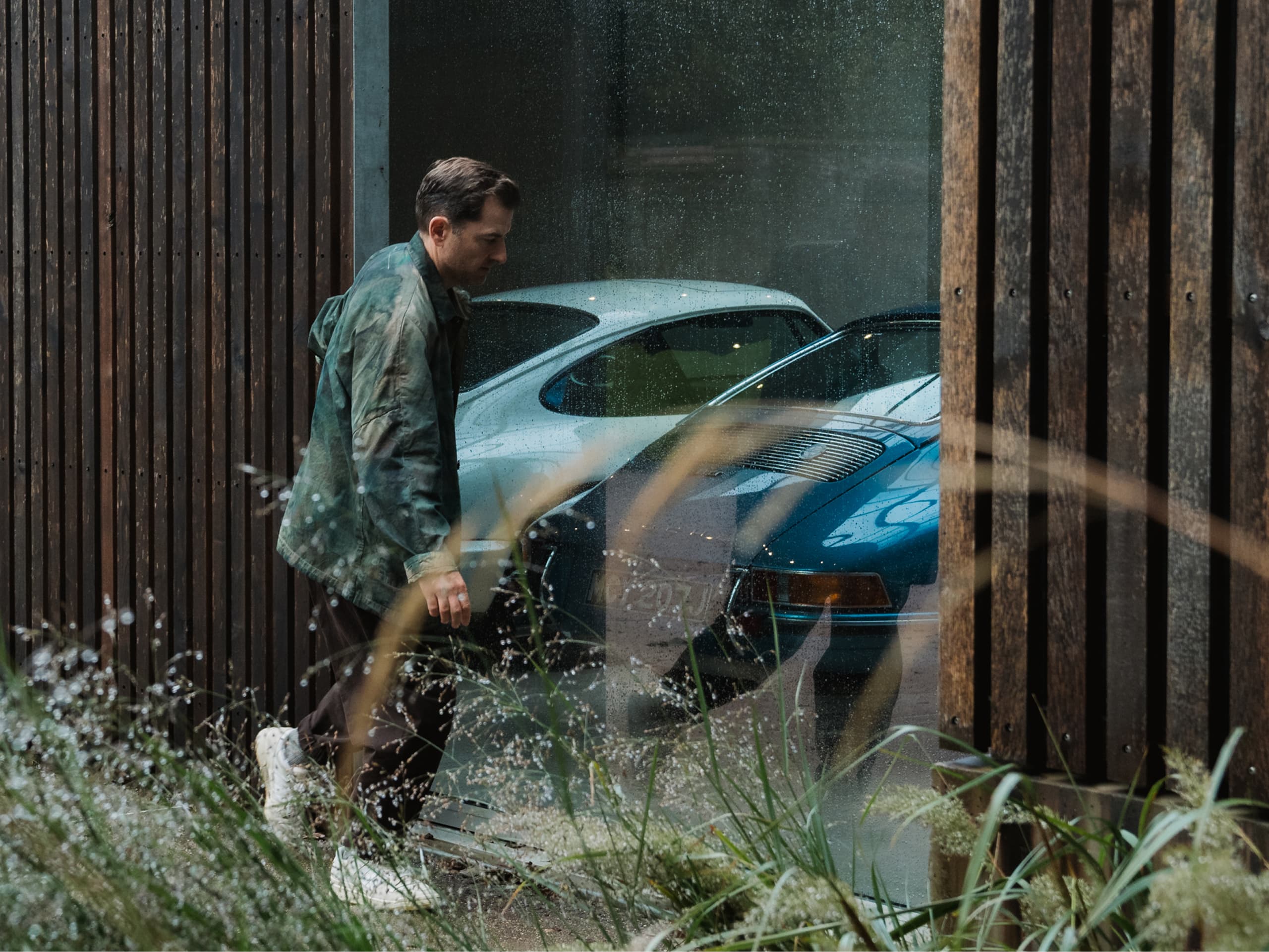 Mann in grüner Jacke geht an einem Fenster vorbei, durch das zwei klassische Autos, ein weißes und ein blaues, durch regengesprenkeltes Glas sichtbar sind.