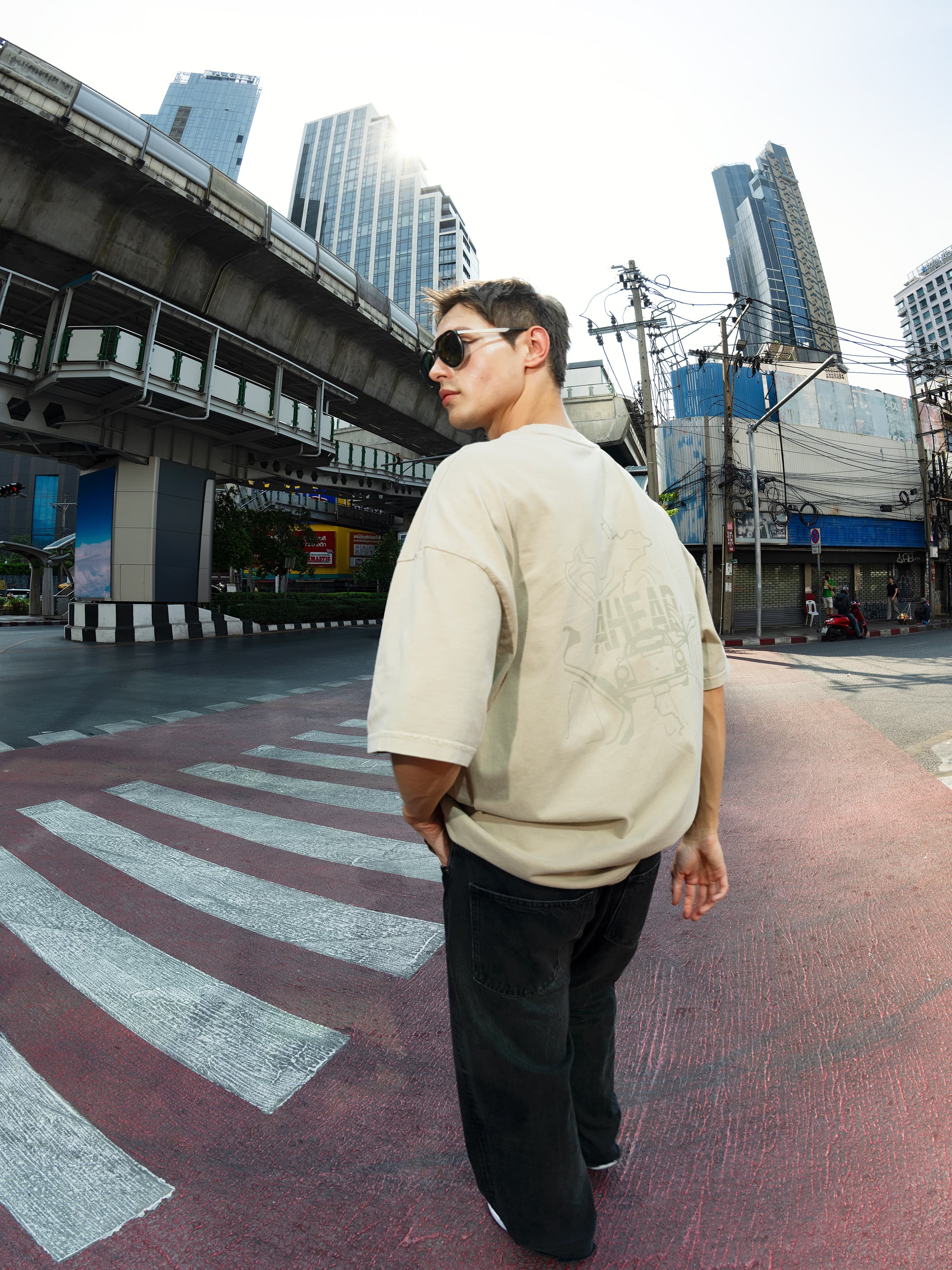Man in beige T-shirt and sunglasses crossing a city street under an overpass, with tall glass buildings behind.