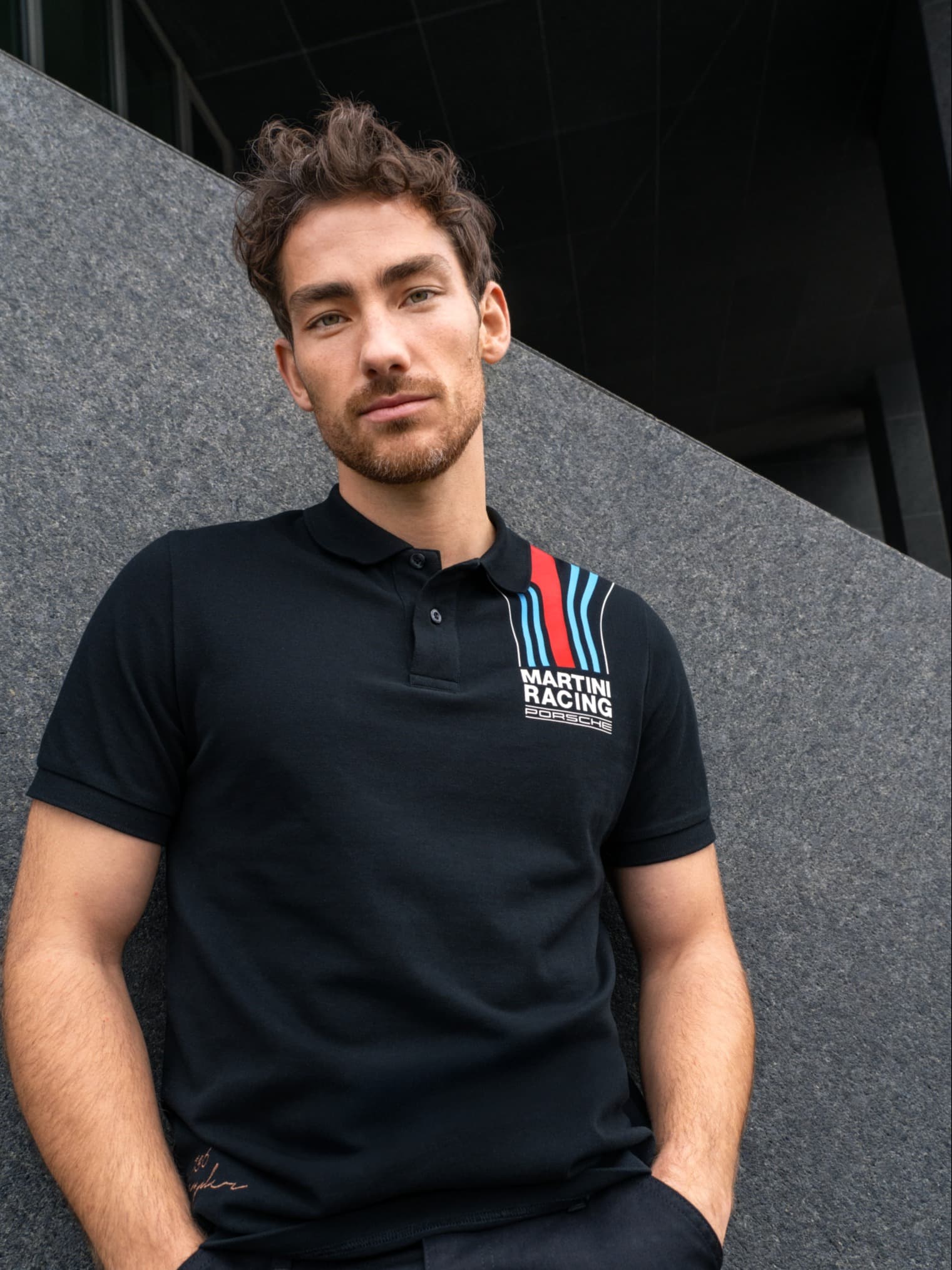 Man in black Porsche Martini Racing polo shirt, standing against a gray wall, looking at the camera.