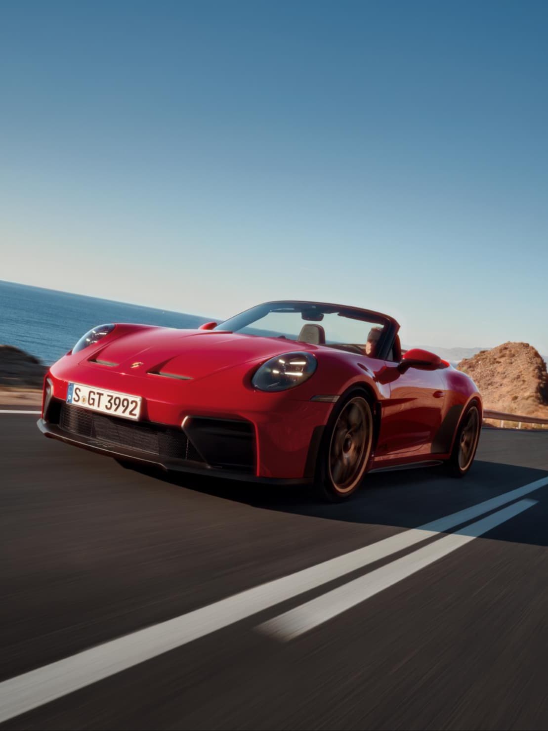 Röd Porsche 911 Cabriolet som kör på en kustväg, havet till vänster och klippor till höger under en blå himmel.