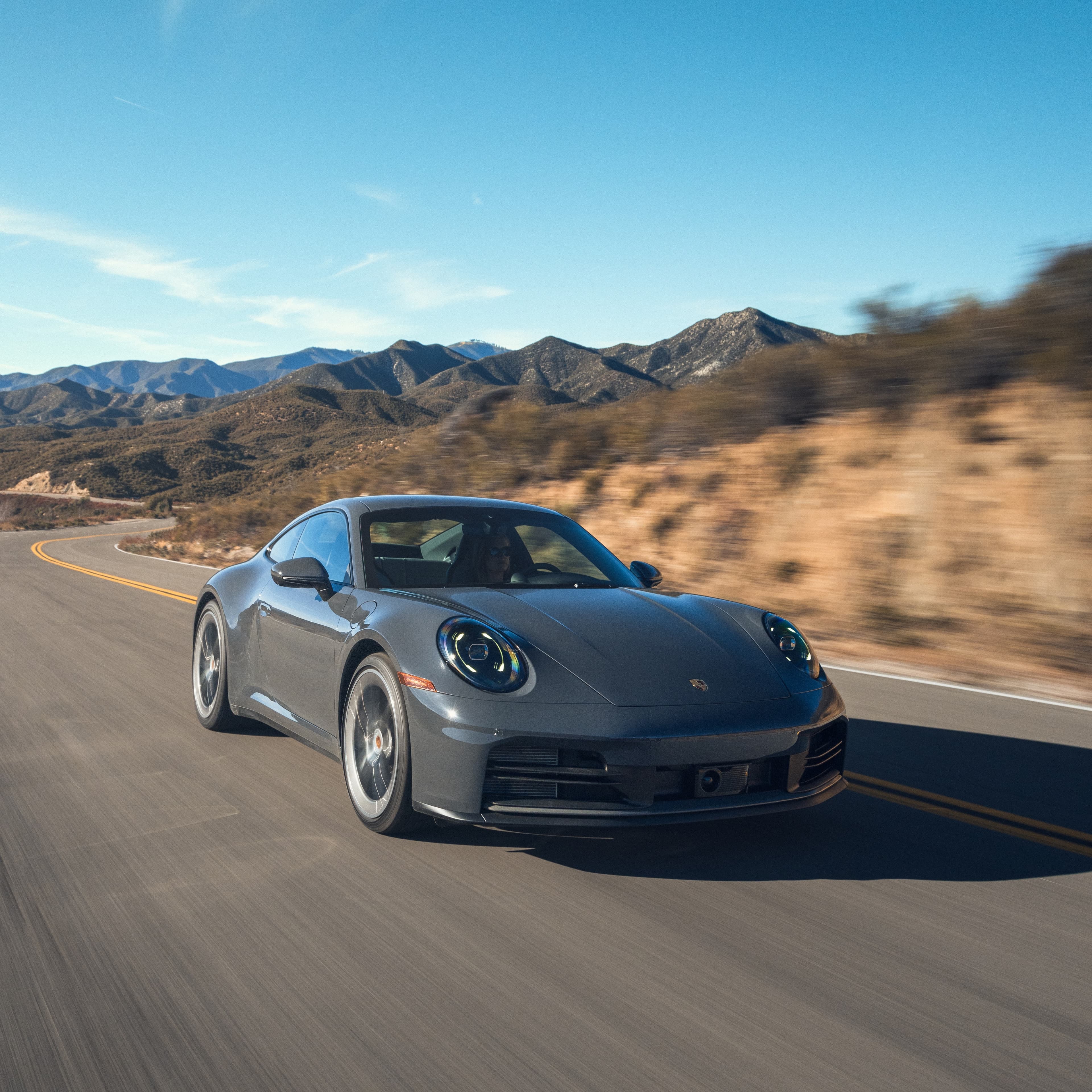Porsche 911 gris roulant sur une route de campagne sinueuse, montagnes et ciel bleu en arrière-plan.