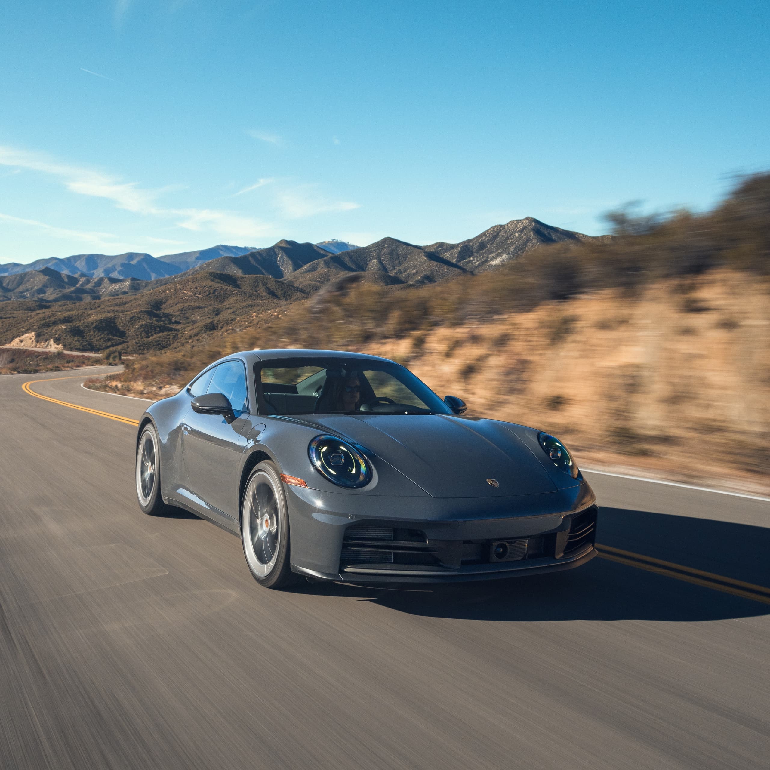 Gray Porsche 911 driving on a winding country road, with mountains and blue sky in the background.