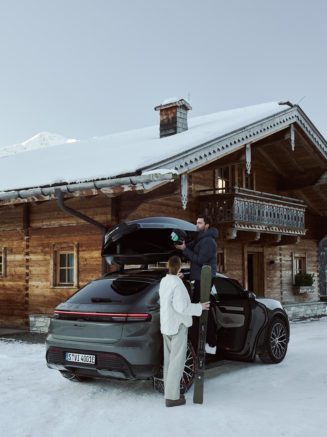 Couple with skis by a black Porsche Taycan in snowy cabin setting, man loading roof box, winter scene.