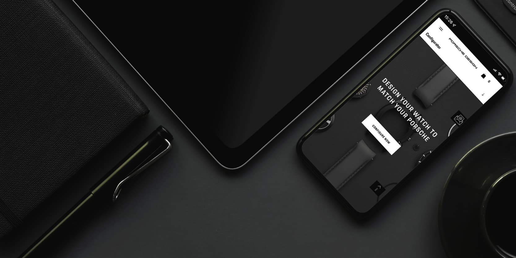 Smartphone showing Porsche Design watch configurator on a dark desk with notebook, pen, tablet and coffee cup