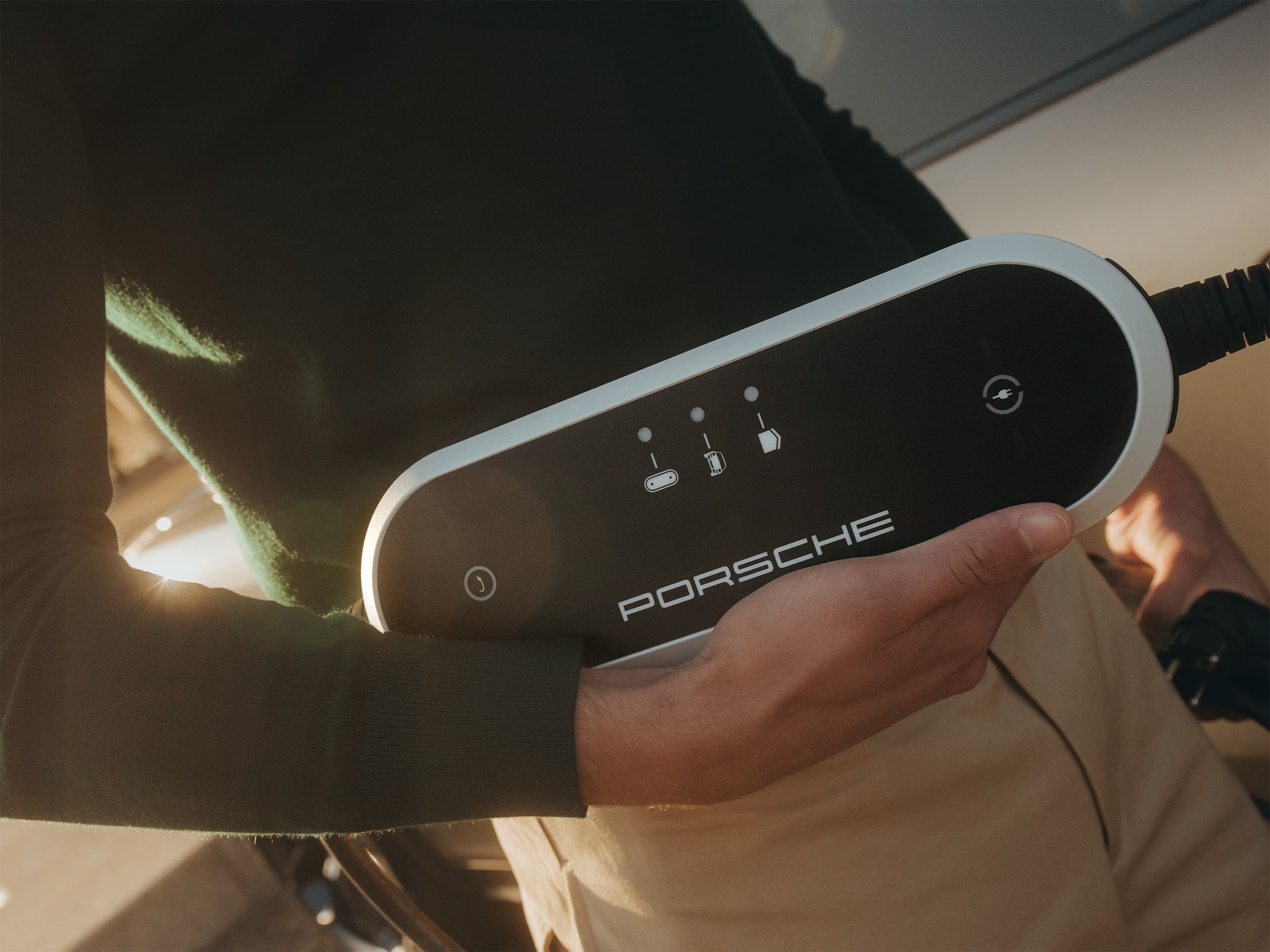Personne tenant un dispositif de charge Porsche contre une voiture, portant un pull vert foncé et un pantalon beige, sous le soleil.