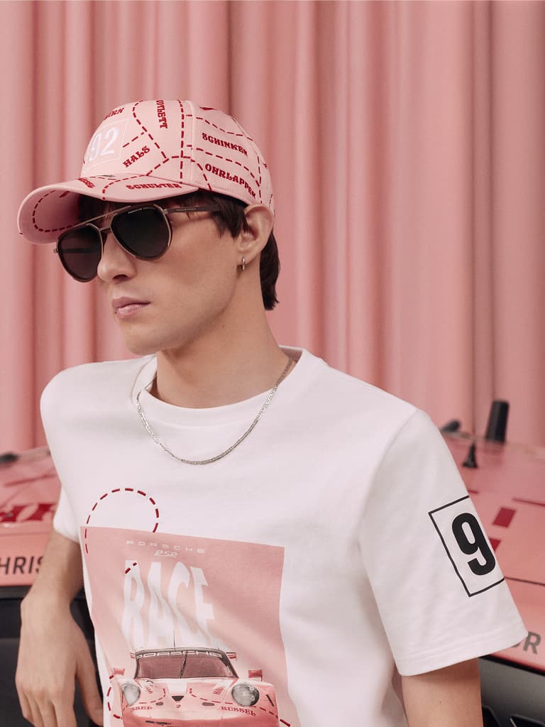 Uomo con cappellino rosa e occhiali da sole, indossa una t-shirt bianca Porsche con grafica di un'auto, su sfondo rosa.