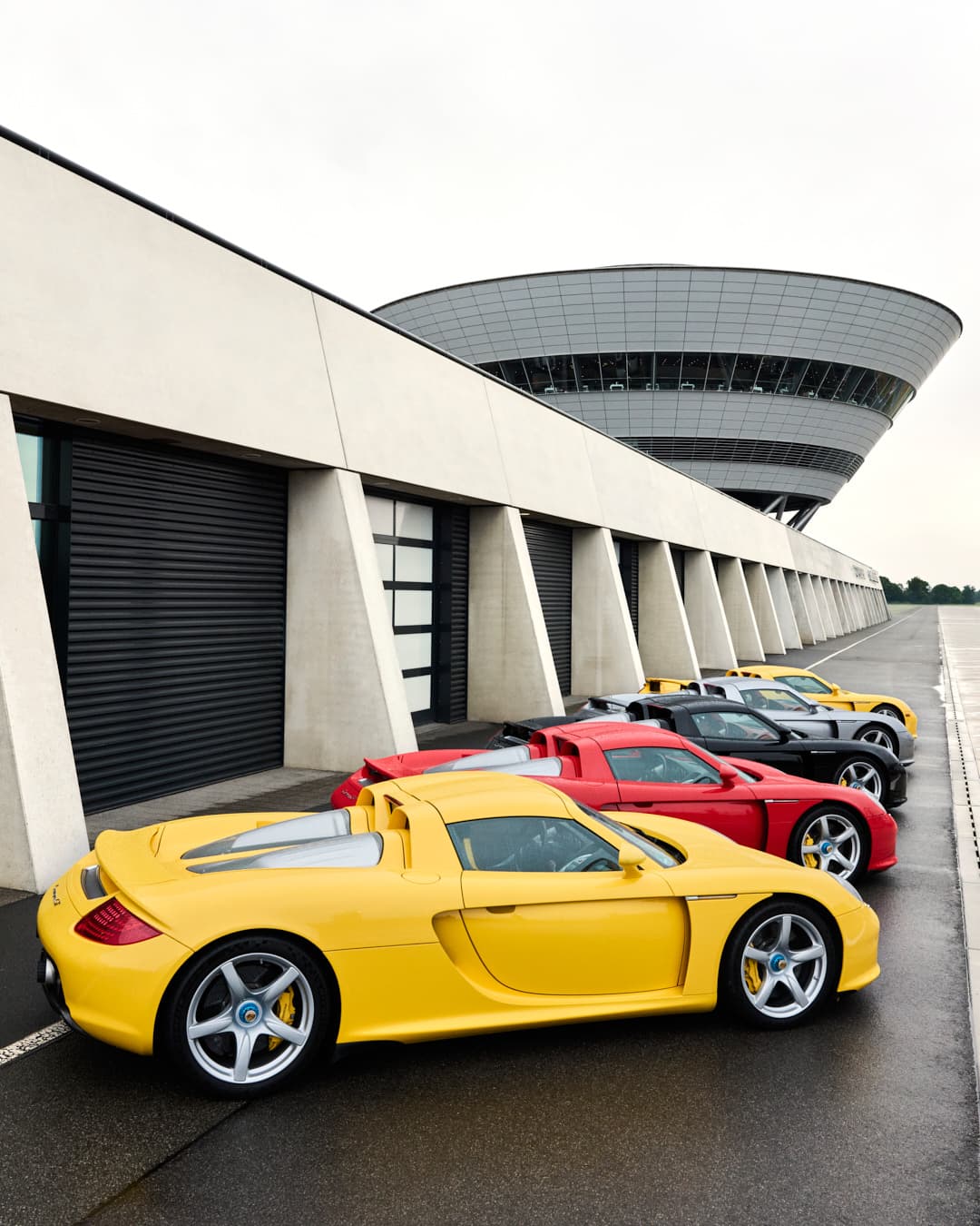 Row of five Porsche Carrera GT vehicles in yellow, red, black, and silver in front of garages