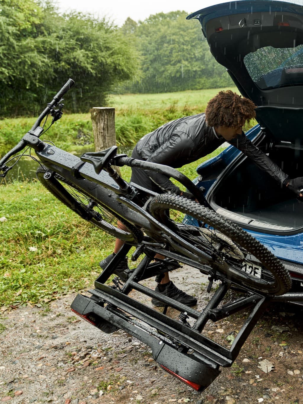Person loading a black bike onto a car rack in the rain, surrounded by greenery.
