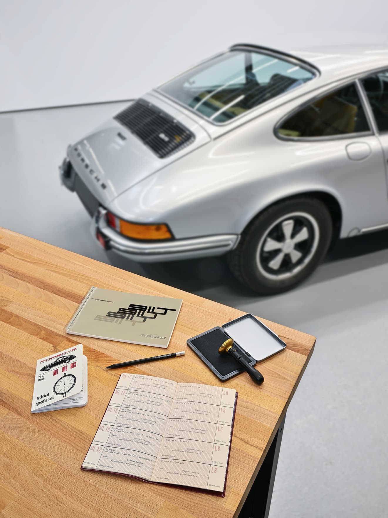 Silver Porsche 911 in garage, wooden table with manuals, magnifying glass, and pencil in foreground.