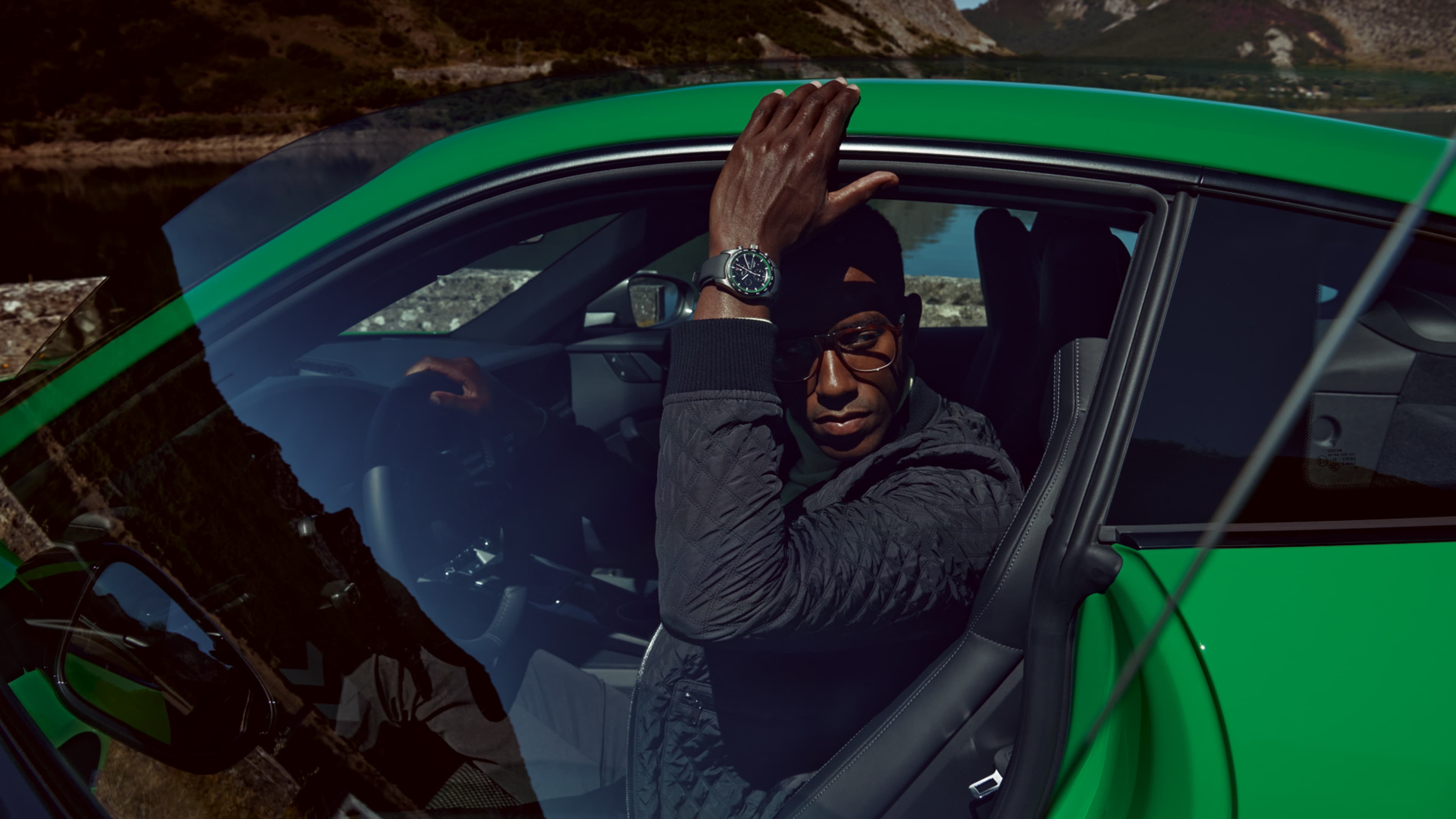 Black Porsche Design chronograph watch with a green leather strap, next to it a steering wheel and a tachymeter ring, with an SUV in the background.