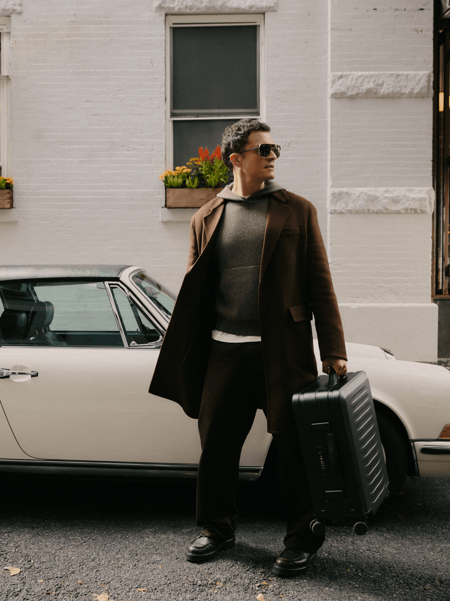 Man in a brown coat wearing sunglasses stands beside a white convertible, holding a black hard-shell suitcase in front of a house wall.