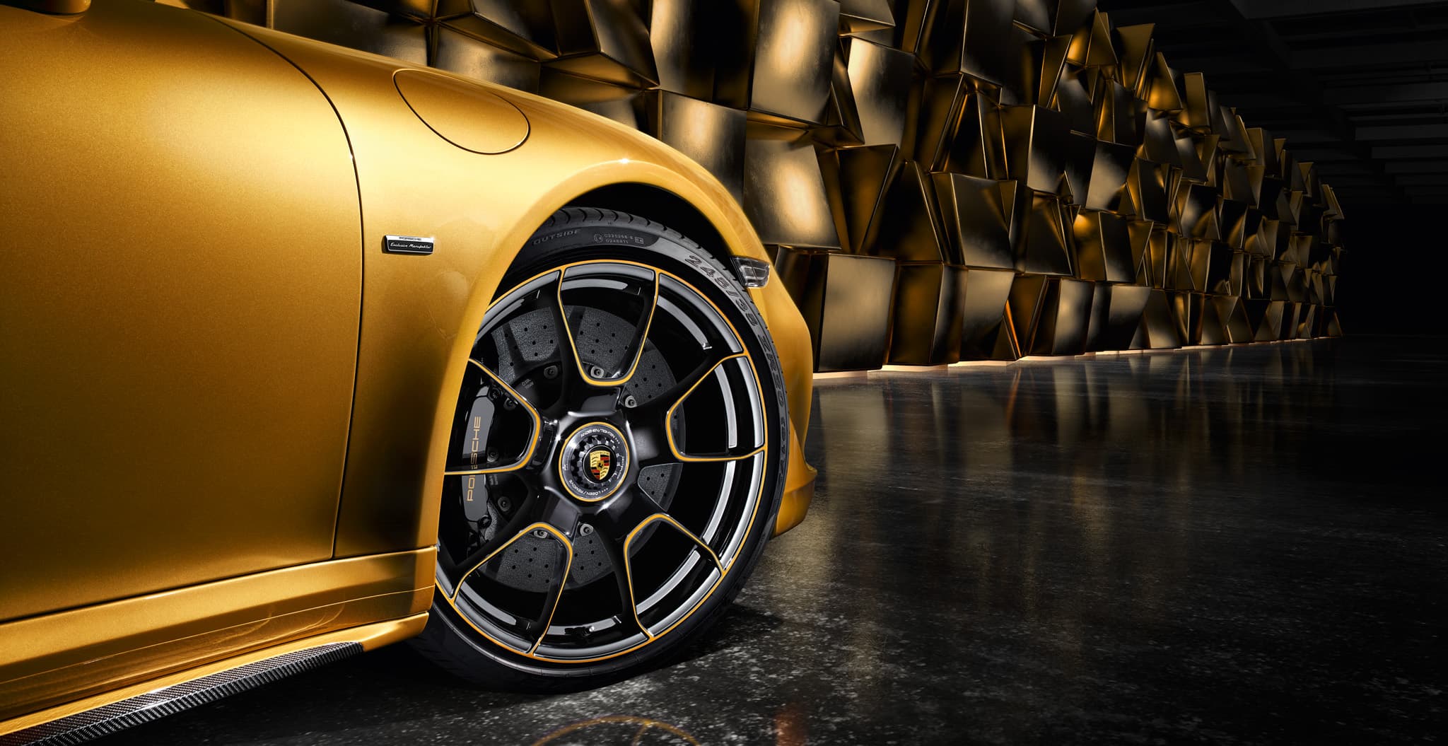 Close-up of gold Porsche front fender and black alloy wheel with gold accents, against a dark geometric wall