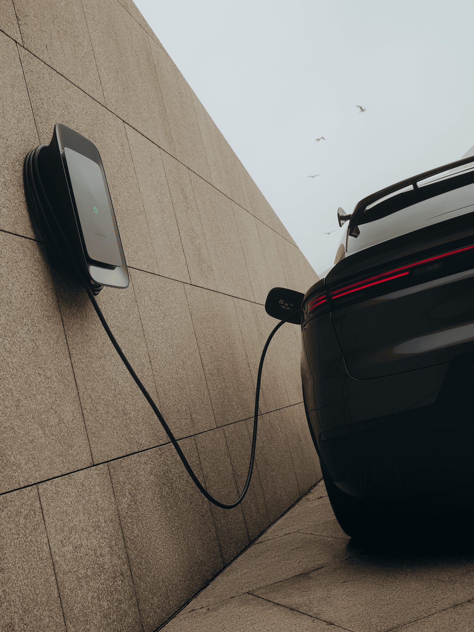 Electric car charging at wall-mounted station, sleek black rear, beige stone wall, overcast sky with birds.