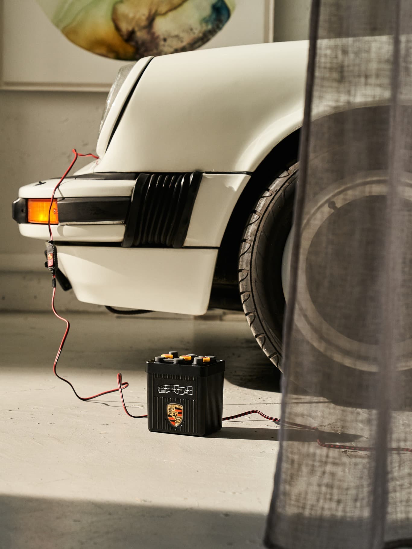 Fehér Porsche autó akkumulátortöltővel csatlakoztatva, garázsban elhelyezve, lágy megvilágítással és részben látható függönnyel.