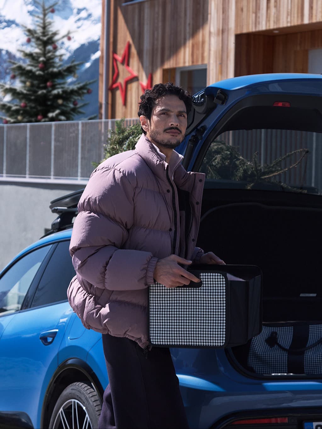 Man in a purple jacket holding a box by a blue car, with a snowy mountain and decorated tree in the background.