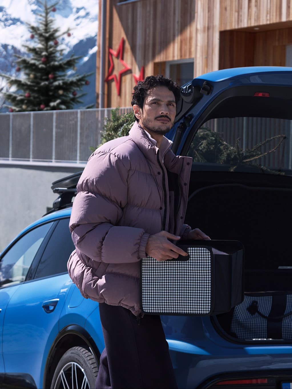 Man in a purple jacket holding a box by a blue car, with a snowy mountain and decorated tree in the background.
