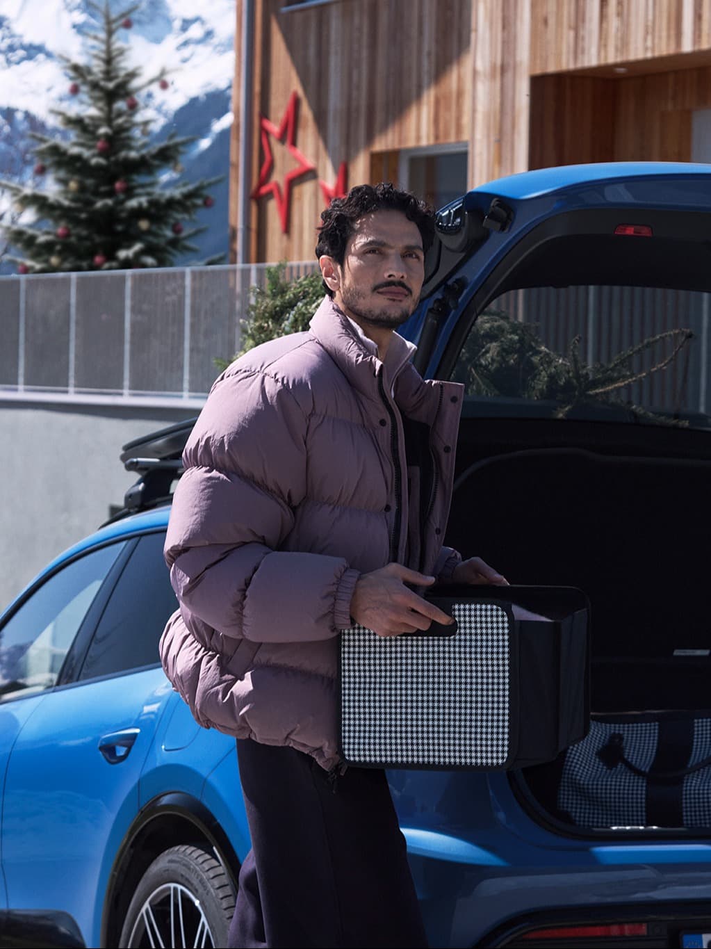 Man in a purple jacket holding a box near a blue car with an open trunk, snowy mountains and wooden house in the background