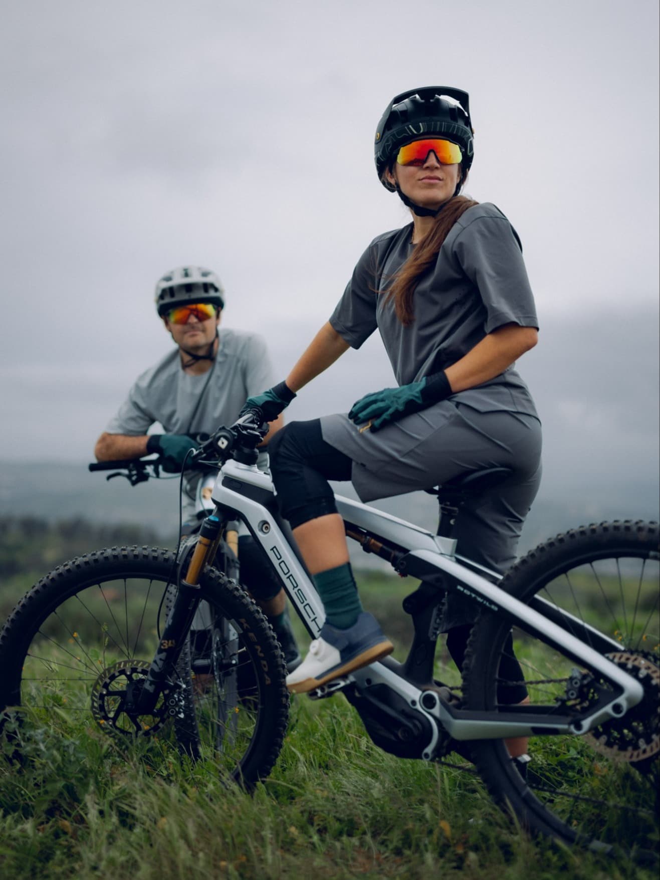 Mountain biker in gray kit with black helmet and mirrored goggles on a Porsche e-bike; second rider behind, cloudy sky.