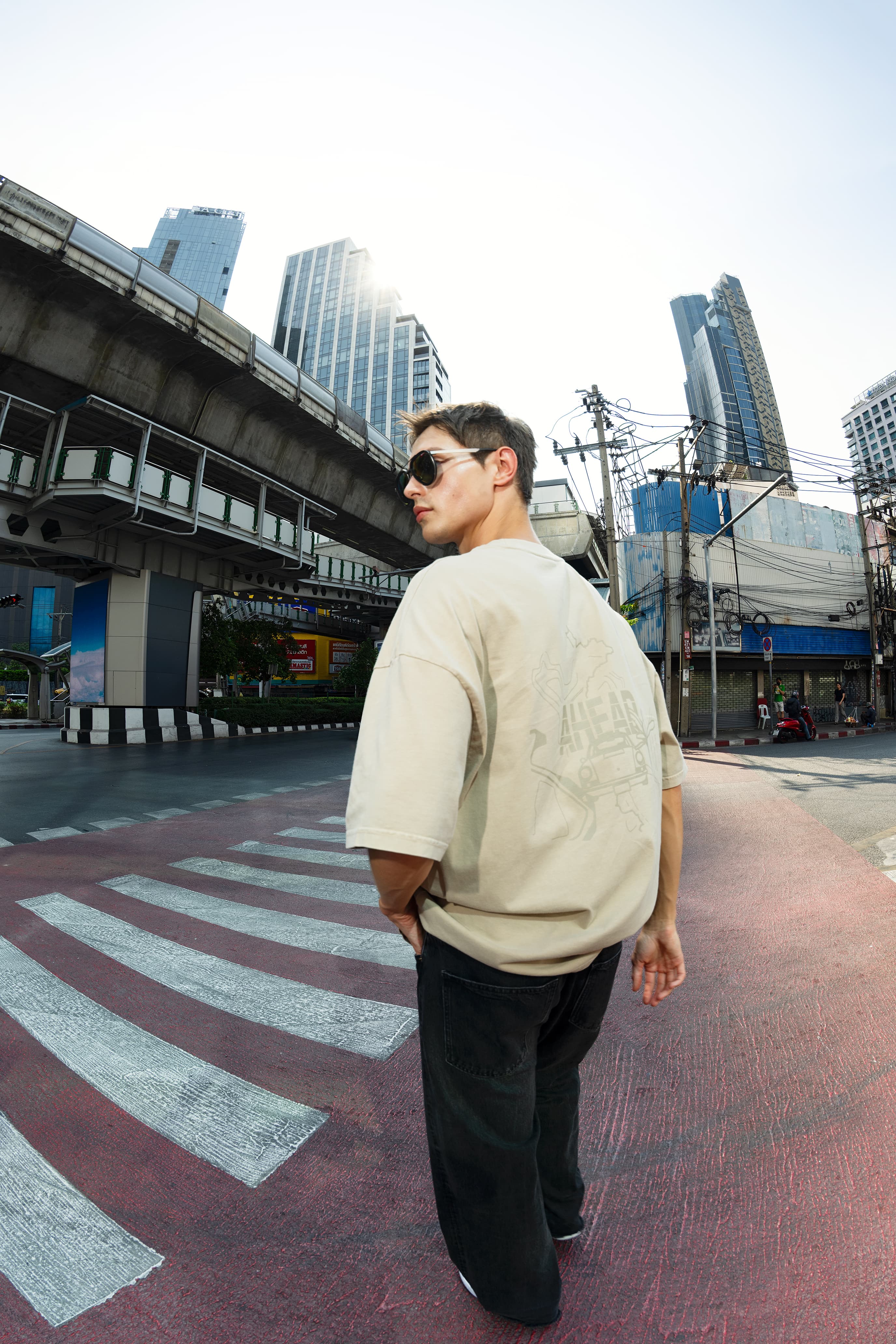 Man wearing sunglasses and a beige T-shirt crossing a zebra crossing, with high-rise buildings and an elevated train in the background.