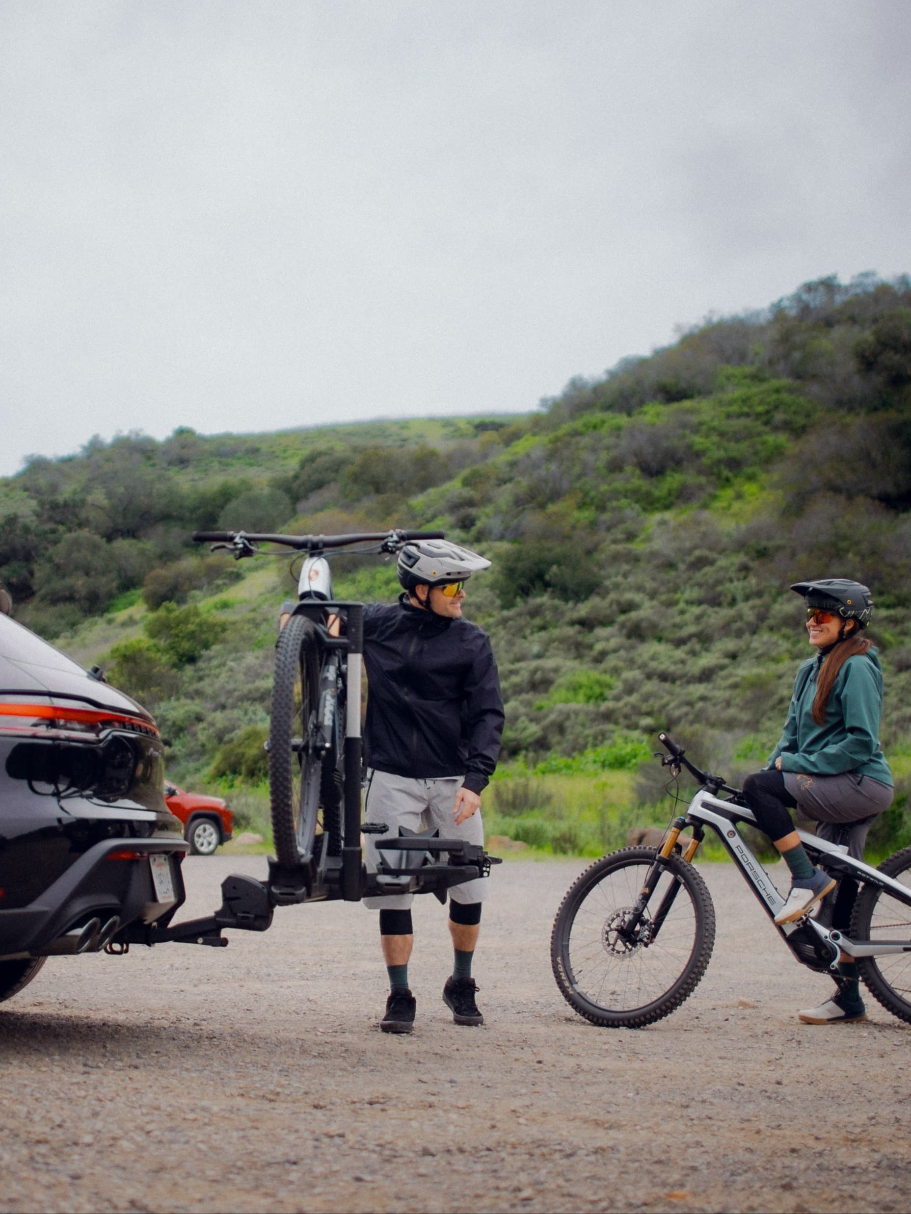 Deux cyclistes casqués près d’une voiture avec porte-vélos arrière, l’un tenant un VTT, collines verdoyantes en arrière-plan.