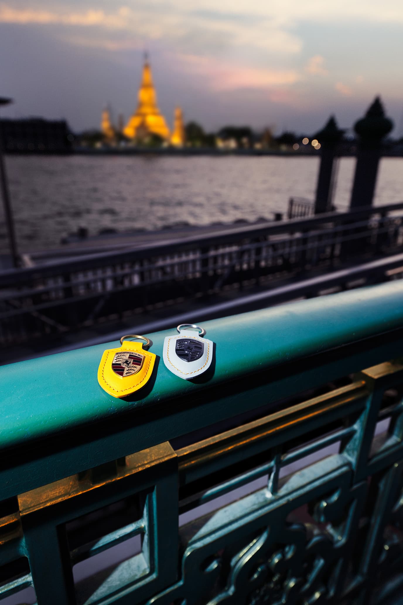 Two leather Porsche key fobs in yellow and white on a green bridge railing, with a river and temple in the background.