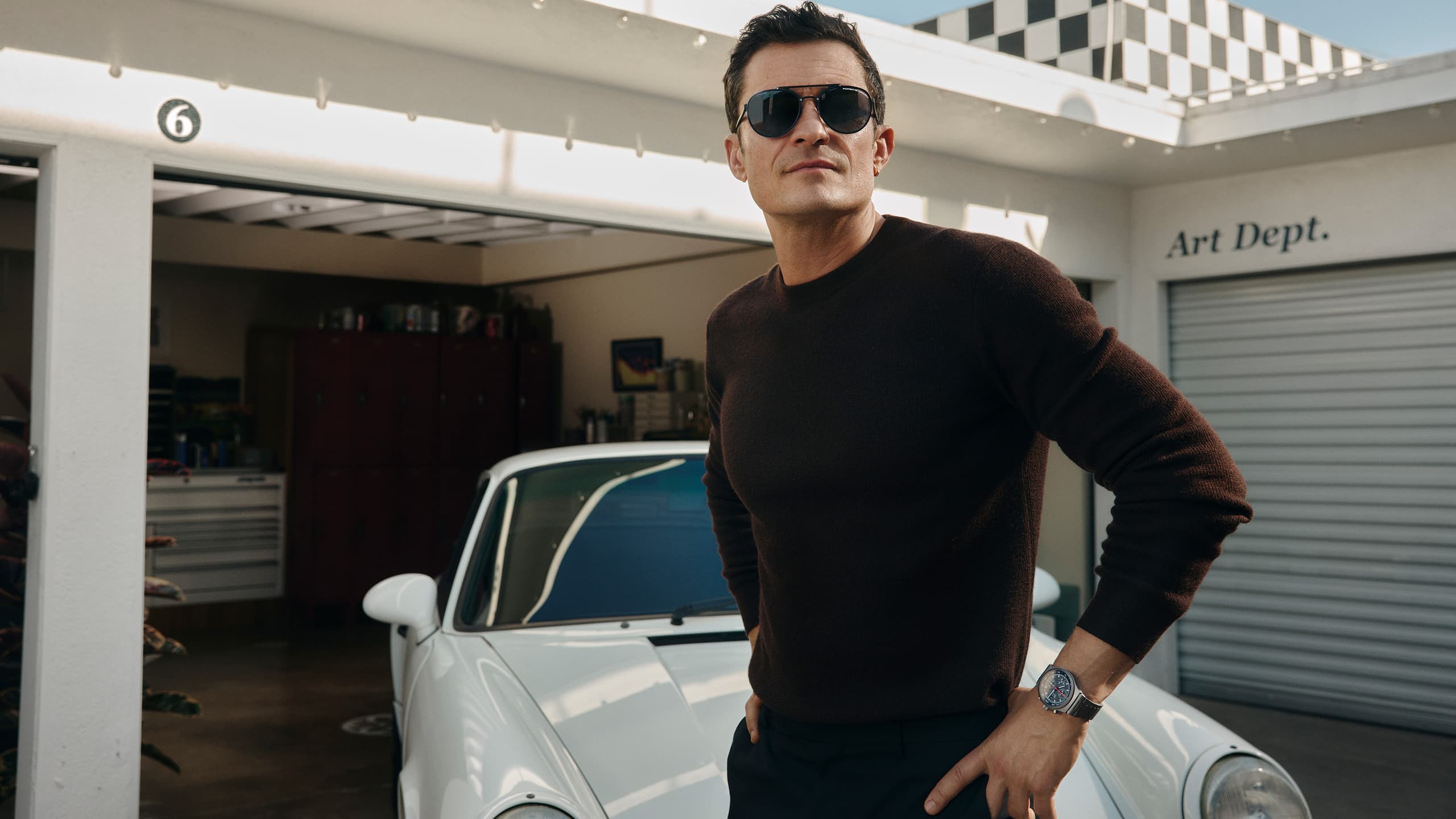 Orlando Bloom wears the Porsche Design Hexagon Eyewear series, standing in front of a garage with a Porsche 911 in the background.