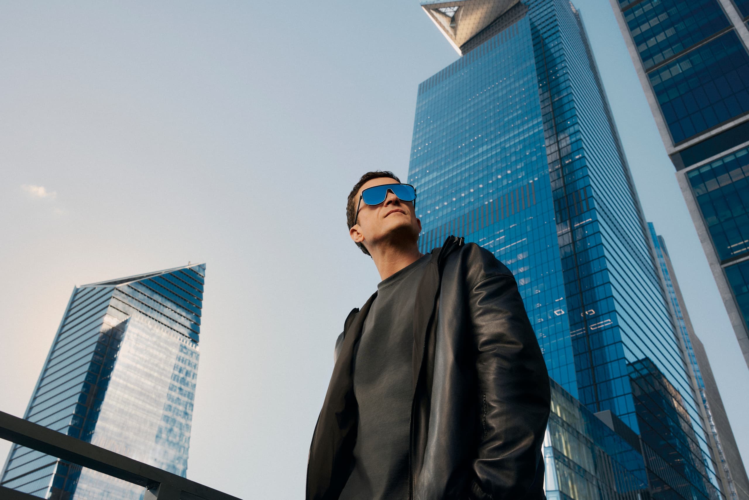 Orlando Bloom wears the Porsche Design Targa #1 Eyewear series, standing in front of a skyline and under a blue sky.
