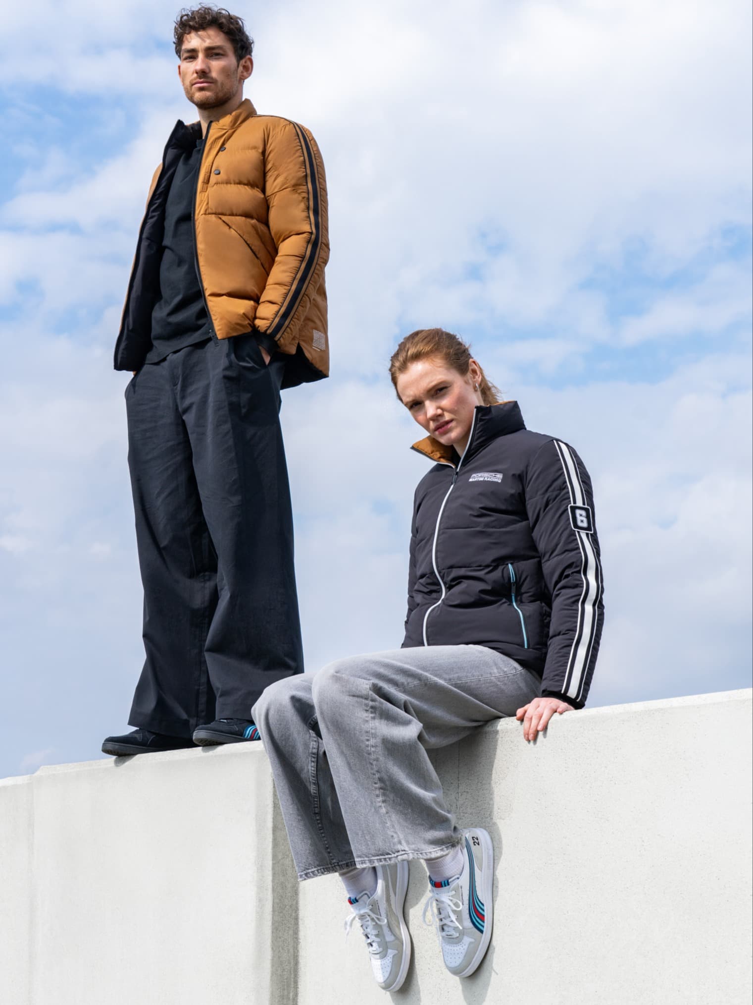 Two people in casual jackets, one brown and one black, stand and sit on a white wall against a cloudy sky.
