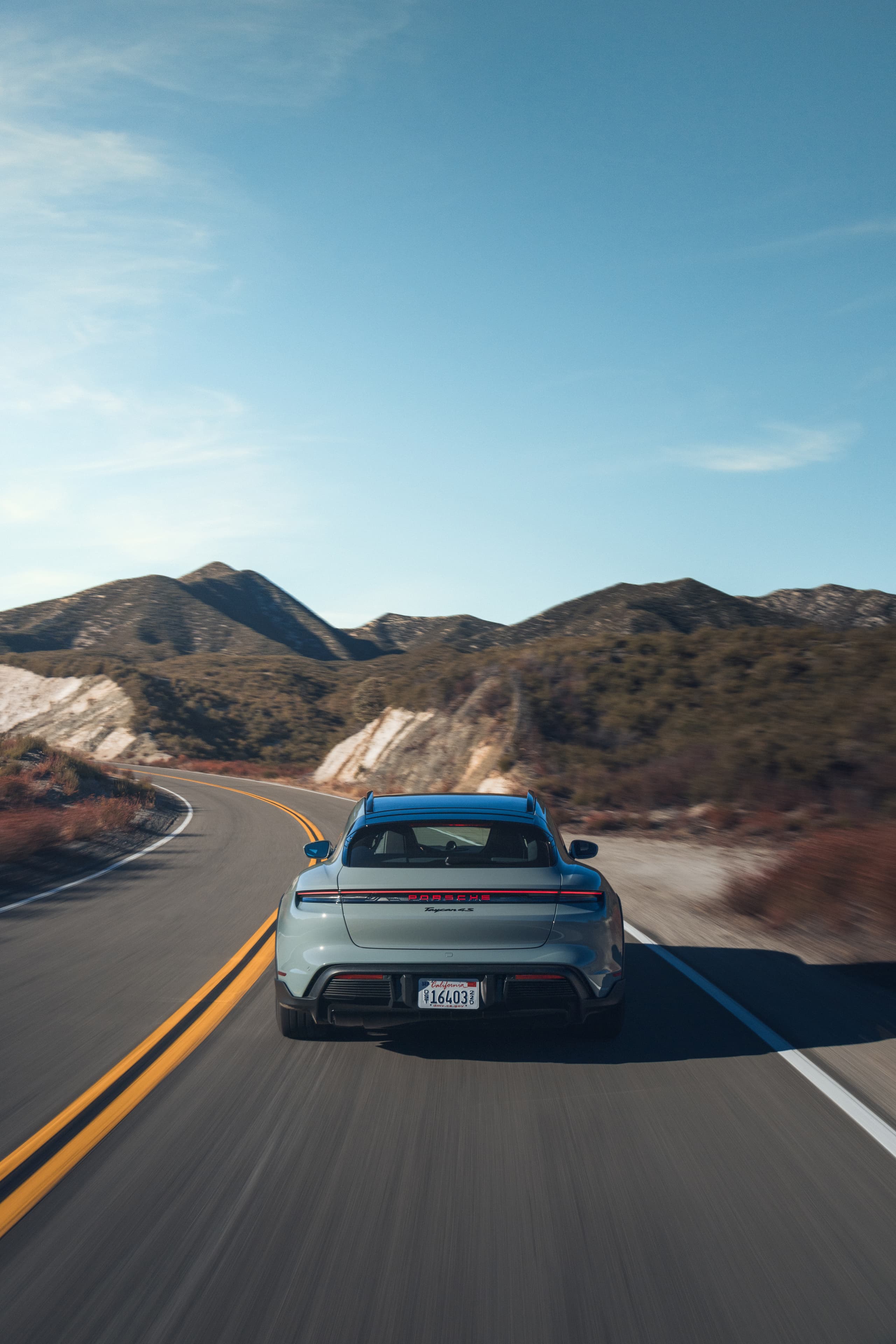 Stříbrný Porsche Taycan jedoucí po klikaté silnici horskou krajinou pod jasnou modrou oblohou.