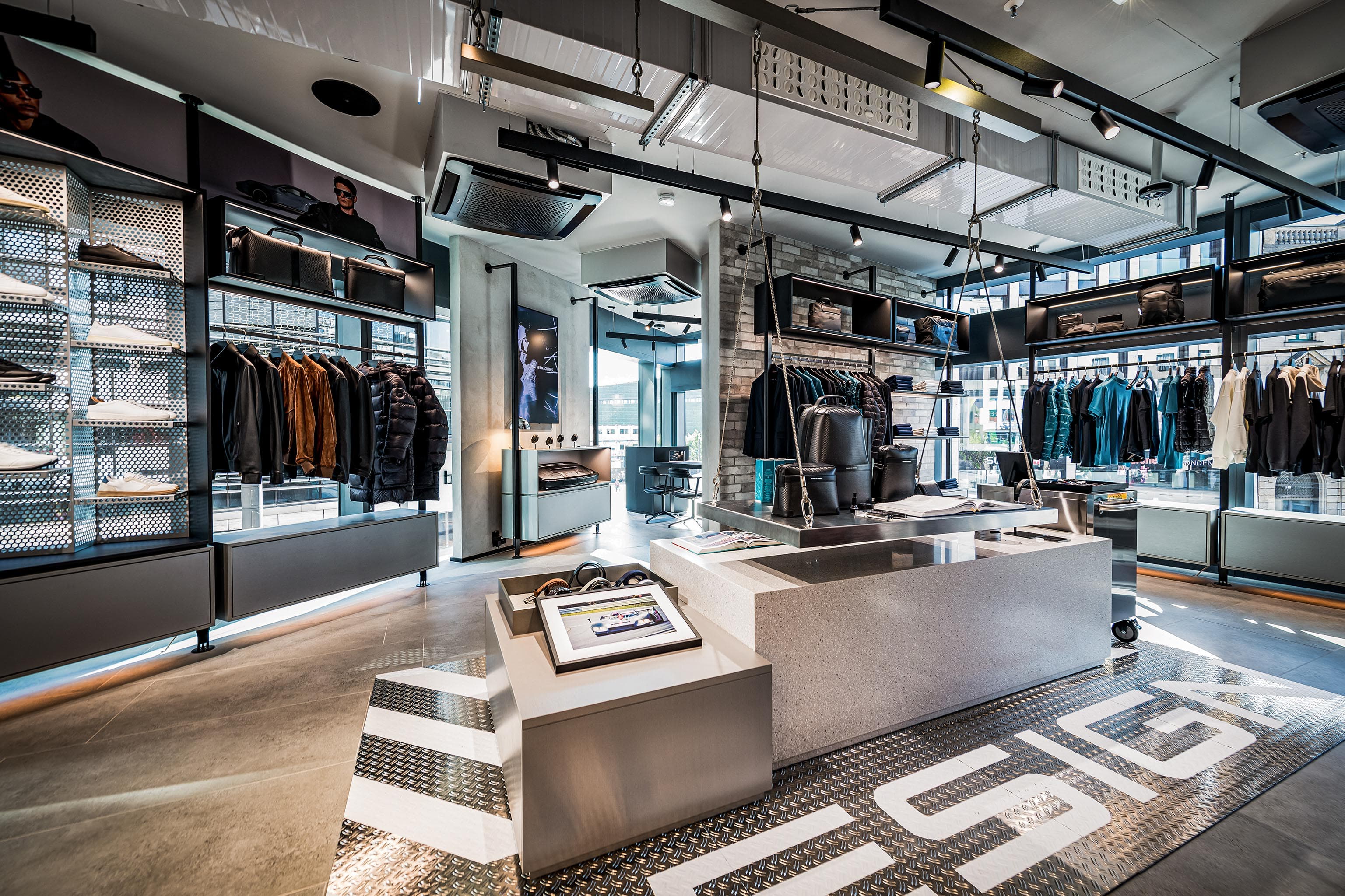 Modern Porsche shop interior with clothing racks, shoes and bags on shelves, and a central display table under spotlights.