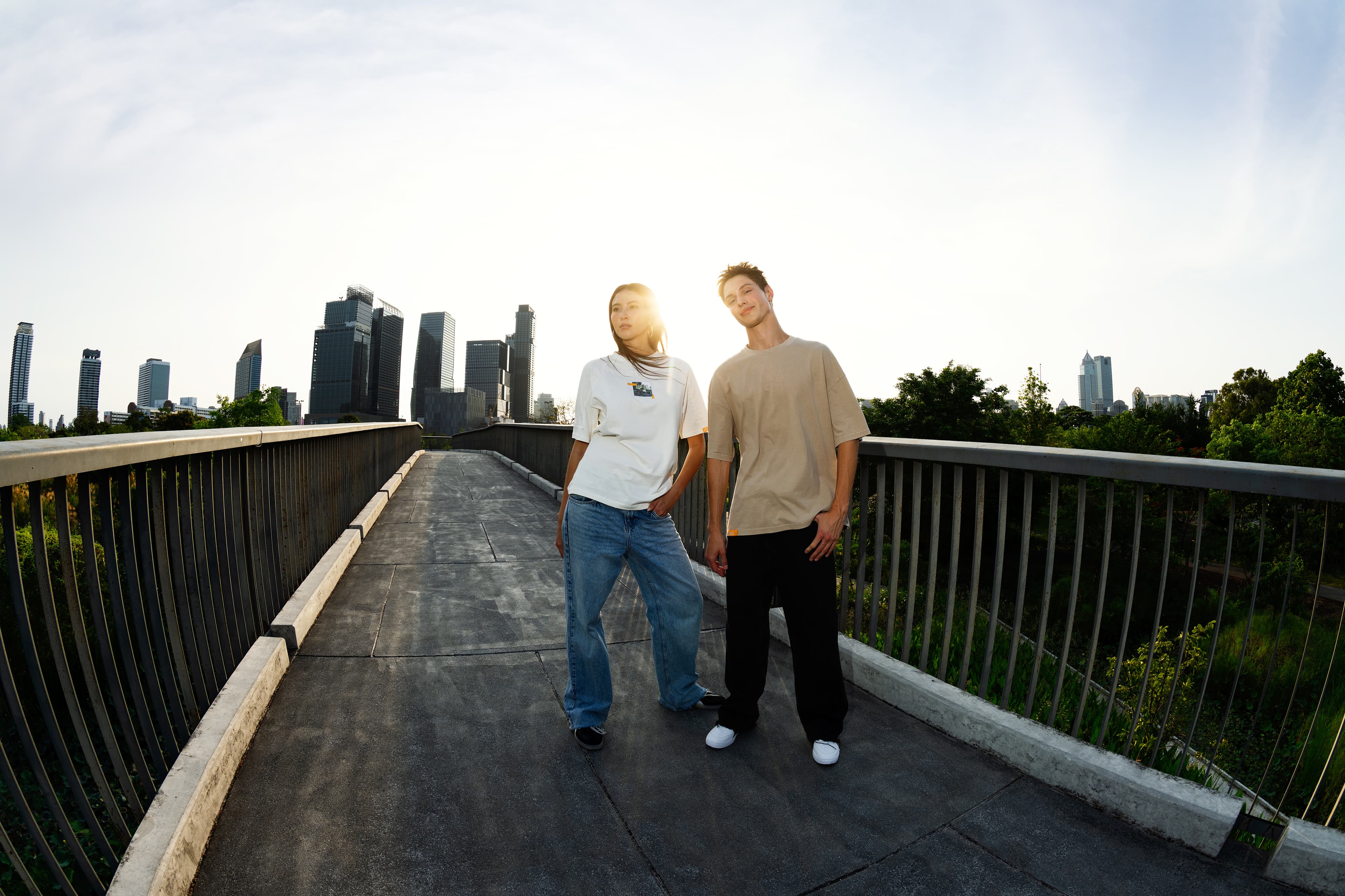 Due modelli su un ponte al tramonto, con T-shirt Porsche AHEAD beige e bianca e jeans, skyline della città sullo sfondo.
