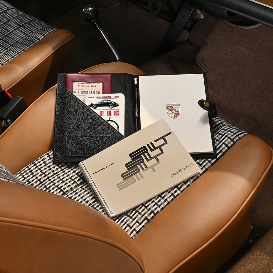 Porsche manuals and documents on a houndstooth car seat, featuring a black leather folder and a white notebook with logo.