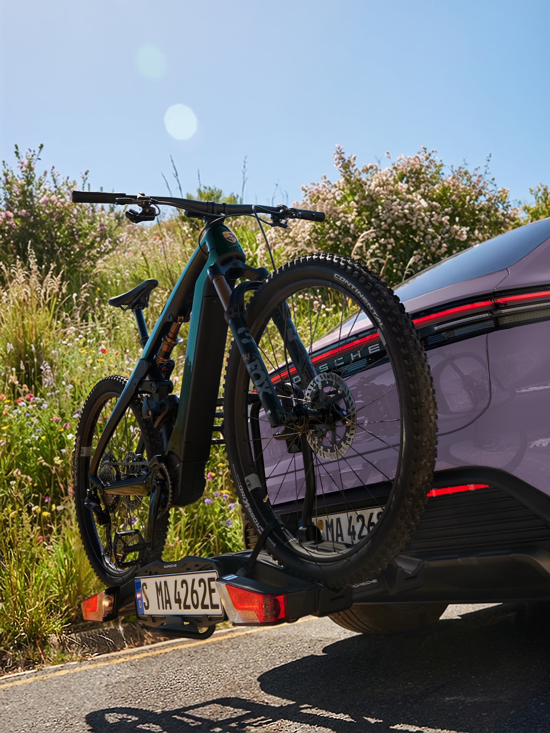 Cykel monterad på en lila Porsche bak, mot en solig bakgrund med grönska och blommor.