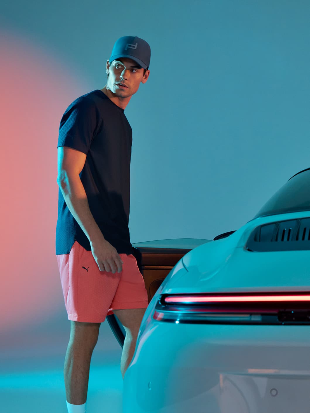 Man wearing a T-shirt, shorts and a Porsche Design Sporswear cap and leaning back against a Porsche