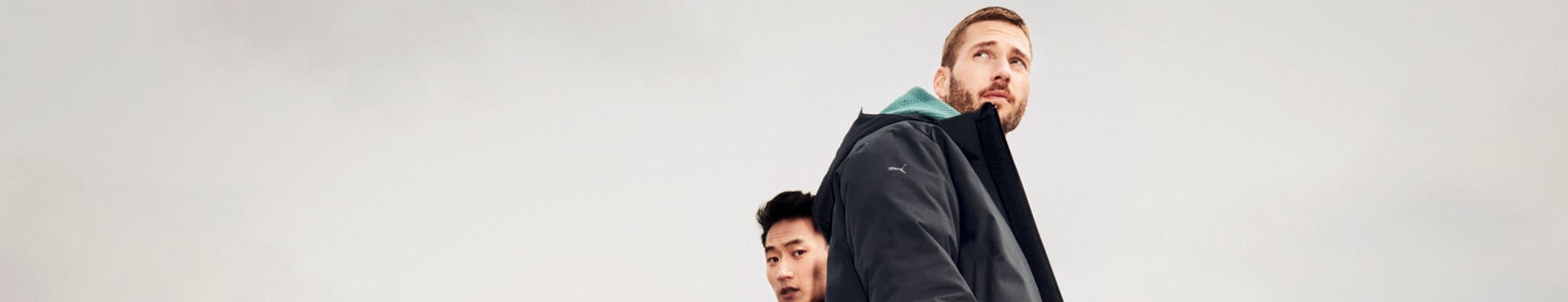 Two men photographed from below are wearing clothes from Porsche Design Fall Winter Sport collection. The man with black hair looks down at the camera, while the man with brunette hair looks into the distance.