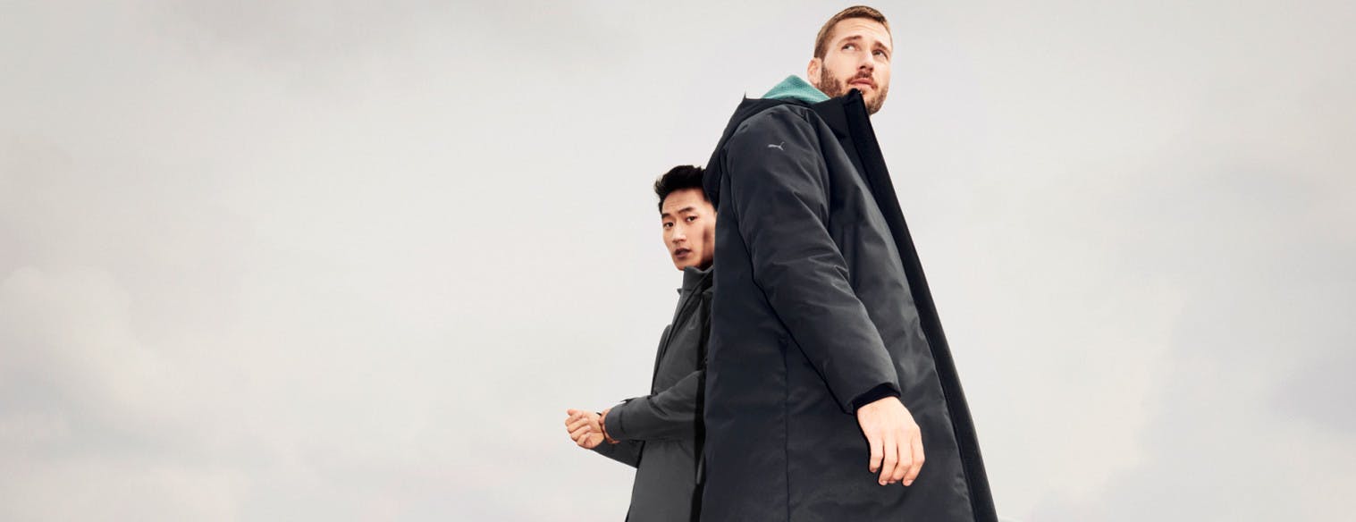Two men photographed from below are wearing clothes from Porsche Design Fall Winter Sport collection. The man with black hair looks down at the camera, while the man with brunette hair looks into the distance.
