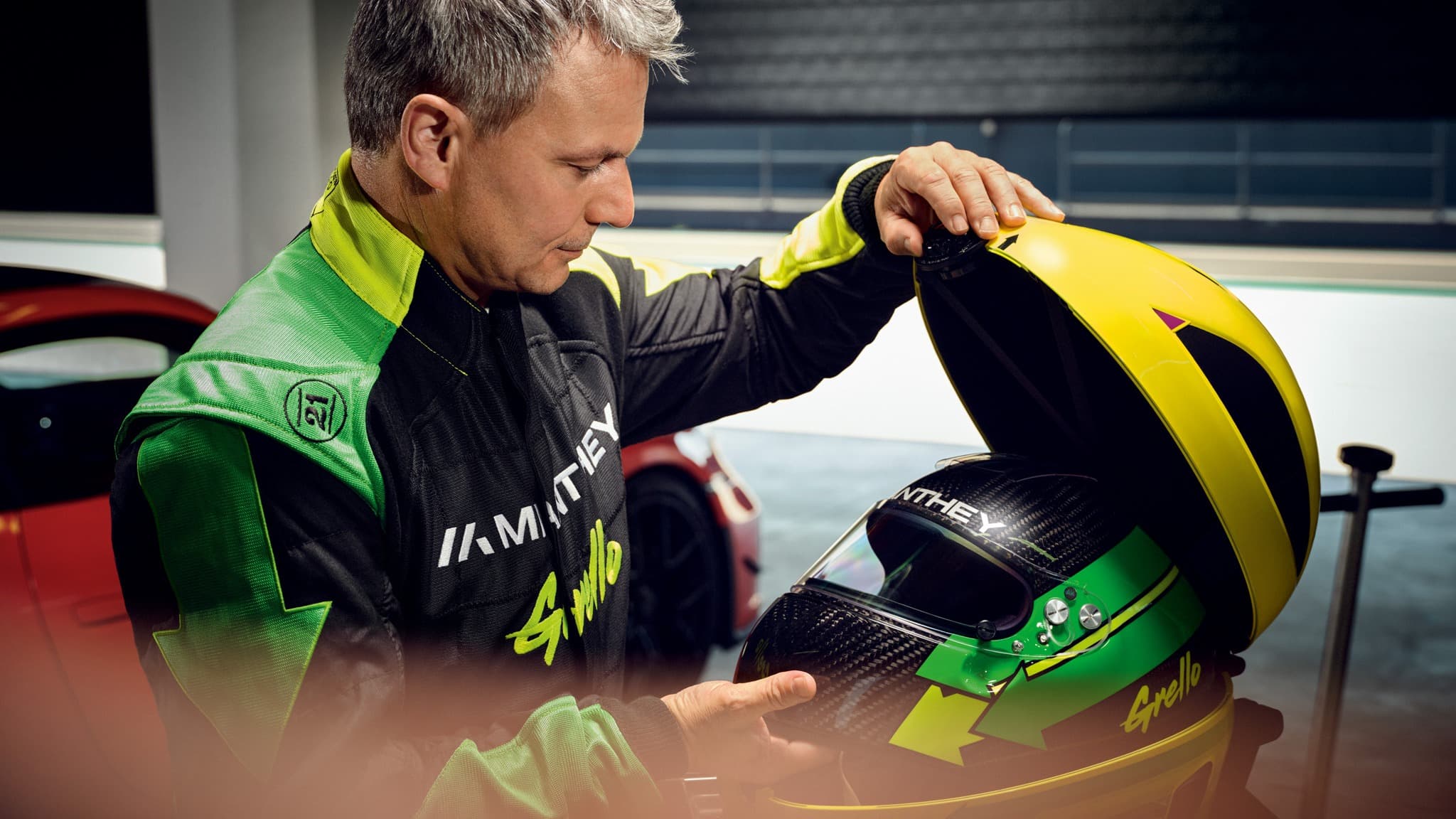Homme en combinaison de course tenant un étui de casque jaune avec un casque vert et noir à l'intérieur, voiture rouge en arrière-plan.