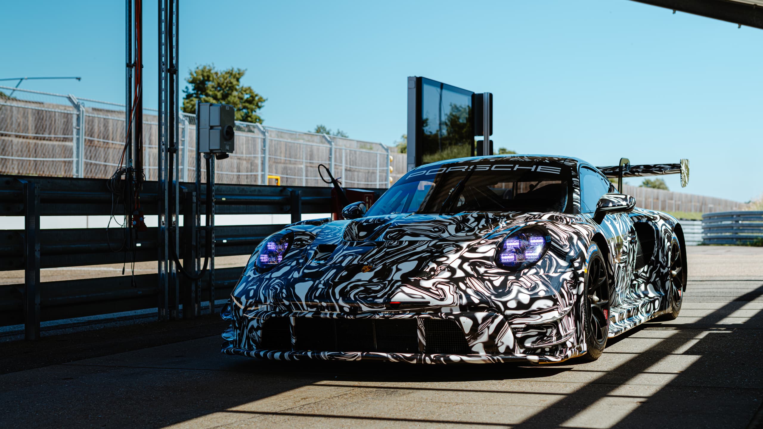 Camouflaged Porsche race car with purple headlights in a pit lane, surrounded by fencing and trees under a clear blue sky.