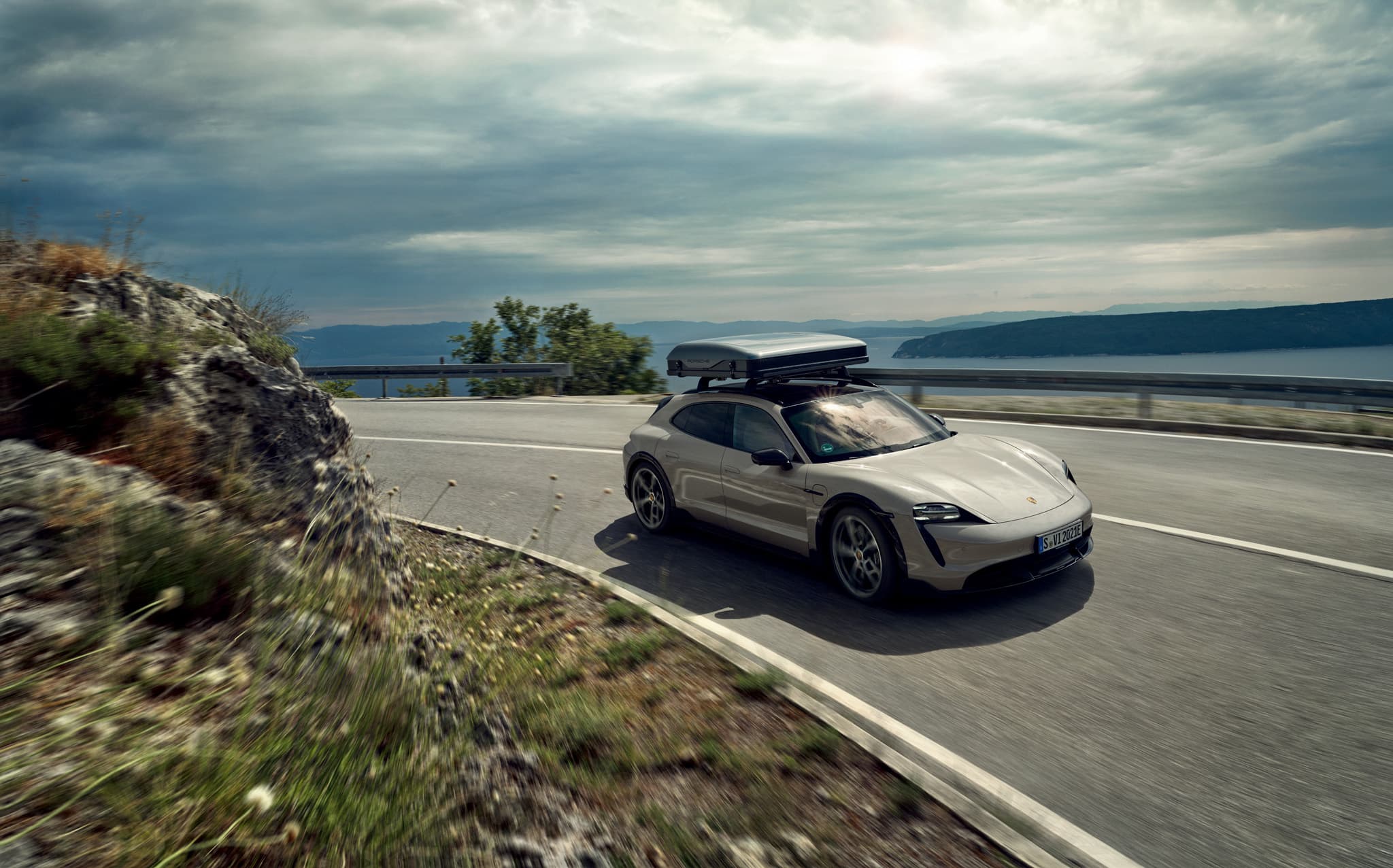 You can see a grey Porsche Taycan with a roofbox driving on a road