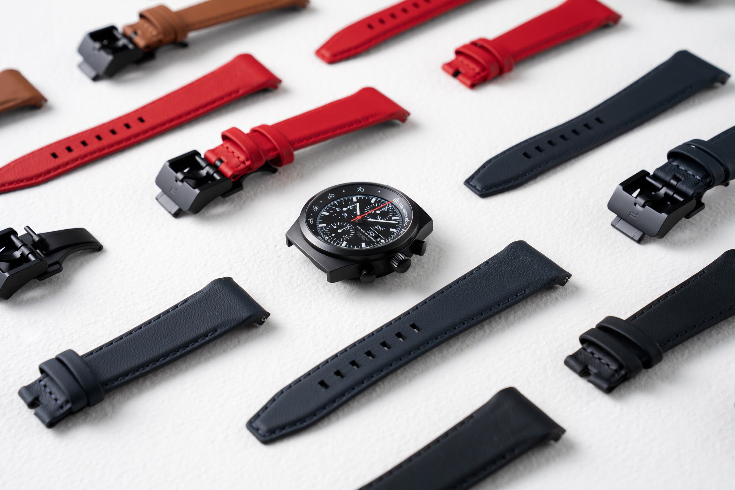 Zwart chronograafhorloge met verwisselbare leren bandjes in rood, zwart en bruin op een lichte achtergrond